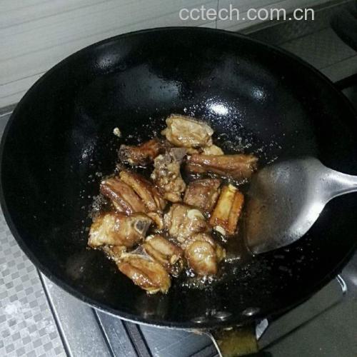一位厨师正在开放式厨房中操作炒锅,左手持锅柄翻炒排骨,右手拿勺准备调味,台面上摆放着切好的菠萝、调料瓶、姜葱等食材,背景为明亮的窗户和绿色植物,体现健康烹饪环境 厨房操作全景图