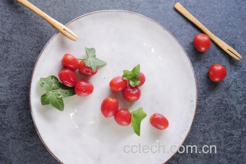 一张俯拍照片,展示一整盘精心摆盘的圣女果夹乌梅,每颗红润饱满的果子中间都嵌着一段乌梅,表面泛着淡淡光泽,撒有白芝麻和绿色薄荷叶点缀,盛放在浅灰色石质餐盘中,背景模糊,突出食物主体 一盘摆盘精美的圣女果夹乌梅,点缀芝麻与薄荷叶