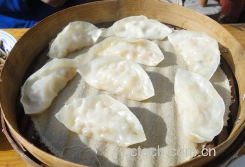 全景摆盘:20个饱满元宝形饺子整齐码放于青花瓷盘,皮色透亮微黄,隐约可见内里翠绿馅心,撒少量白芝麻与嫩芹菜叶点缀,背景为木质料理台与西葫芦芹菜新鲜食材 西葫芦芹菜饺子成品摆盘图