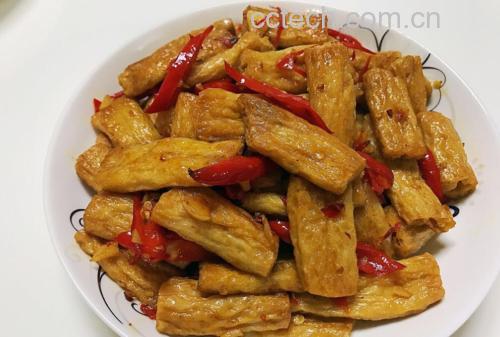一盘金黄色的豆腐干与翠绿色尖椒交错翻炒在黑色铁锅中,表面微焦泛光,旁边散落着红色干辣椒段和青蒜叶,背景虚化体现厨房实操场景 尖椒炒豆腐干特写