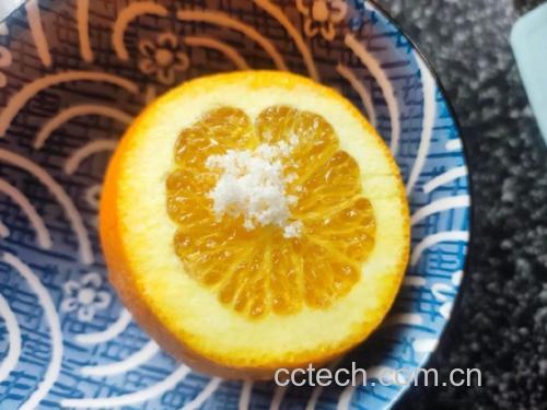 一只手正将切开的橙子放入白色瓷碗中,橙子切面向上,另一只手捏着一小撮食盐准备撒下,桌面上有盐罐、小勺和擦干的橙子,光线柔和,突出操作细节 盐蒸香橙装碗过程