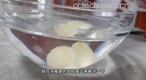 一个黑色冻梨浸泡在透明玻璃碗的清水中,水面微微泛起气泡,冻梨表面有细微冰晶融化痕迹,背景虚化的厨房环境显示为冬季室内,窗外可见薄雪,强调‘冷水缓化’的真实场景 冻梨解冻过程特写