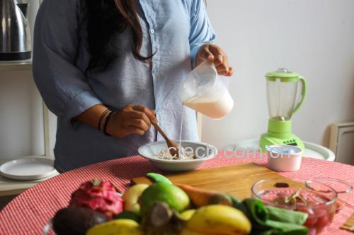 一位中年女性在明亮现代的厨房中准备早餐,桌上有一碗燕麦粥、一杯温水和一小碟切好的紫皮蒜片;她正用小勺将蒜泥加入水中,动作自然,窗外阳光洒入,营造出健康生活氛围 家庭厨房中食用紫皮蒜场景