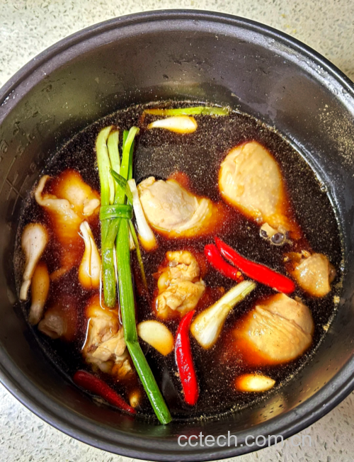 铸铁锅中正在炖煮的鸡腿肉,汤汁微沸,周围环绕着葱段和姜片,鸡肉呈酱红色,蒸汽升腾,展现浓郁的烹饪氛围 鸡腿肉与葱在锅中炖煮过程
