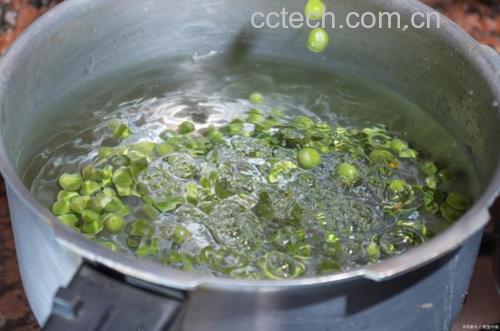 一名厨师正在将新鲜青豌豆倒入沸水中进行焯烫,锅边有蒸汽升腾,旁边放置冰水盆用于后续冷却,操作台整洁,背景为现代厨房环境 青豌豆焯水过程示意图