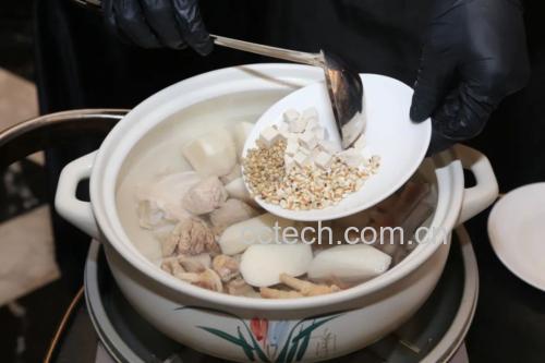 一位厨师在明亮整洁的现代厨房中,身穿白色围裙,正在用木勺搅拌一口黑色砂锅中的汤品,锅中可见鸡块和药材,蒸汽袅袅升起,台面上摆放着三七粉瓶、枸杞罐、红枣和姜片,整体呈现专业而温馨的烹饪氛围 厨房操作场景