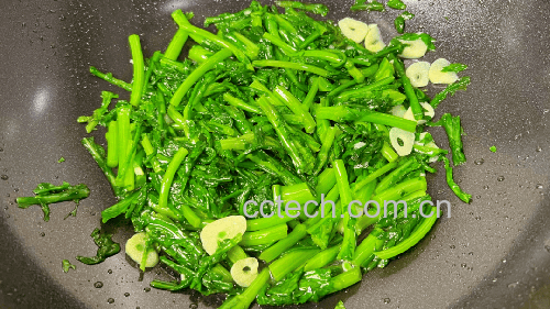 一口黑色不粘锅中,金黄色的蒜片与灰白色平菇一同翻炒,锅边略带焦香,蒸汽升腾;旁边案板上放有切好的青椒丝和葱段,展现家常健康烹饪场景 家庭厨房中正在翻炒平菇的锅具特写