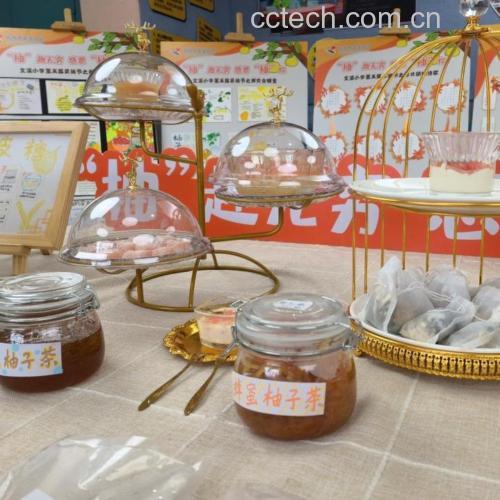玻璃罐中盛装着金黄色的蜂蜜柚子茶,果肉与柚子皮清晰可见,质地浓稠,旁边搭配一个木勺和一杯冲泡好的热饮,背景为厨房操作台 自制蜂蜜柚子茶成品展示