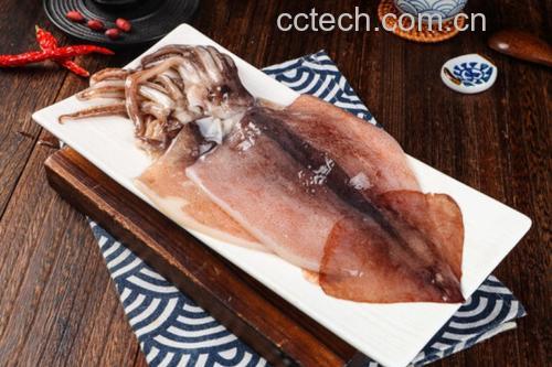 一道刚出锅的小鱿鱼菜肴被盛入白色瓷盘中,鱿鱼呈金黄色微焦状态,卷曲成花状,周围点缀着红色辣椒和绿色葱花,背景为木质餐桌,整体呈现诱人的食欲感 小鱿鱼成品装盘效果