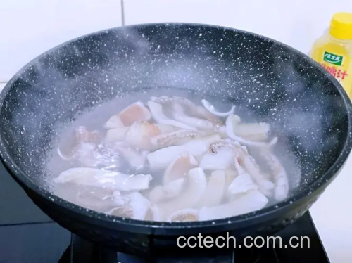 一只手正在将解冻后的鱿鱼放入装有姜片和料酒的沸水中焯烫,锅边有蒸汽升腾,旁边摆放着一碗冰水用于快速降温 图片描述