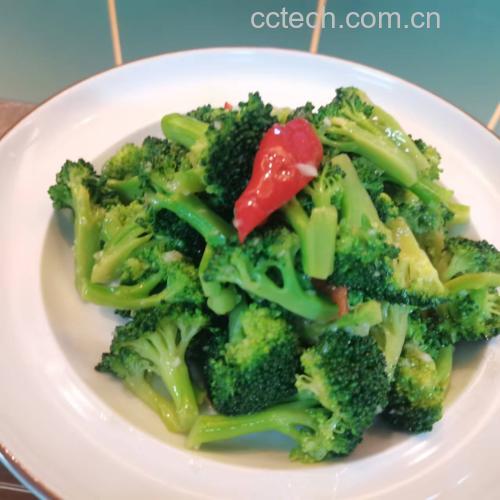 成品凉拌西兰花特写：翠绿饱满的西兰花花球与透亮茎片整齐铺于浅灰陶盘，表面淋有琥珀色酱汁光泽，点缀金黄熟芝麻与细碎小米辣，背景虚化呈现木质餐桌与玻璃水杯
