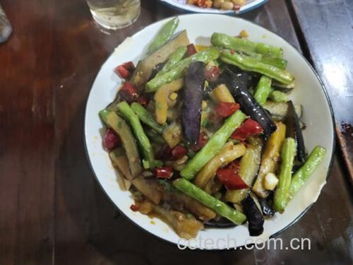 深色炒锅中,茄子条与翠绿毛豆米混合翻炒,汤汁正逐渐收浓,表面泛着油亮光泽,旁边摆放着酱油瓶、蒜末碟等调味容器,背景为家庭厨房环境 锅中正在收汁的毛豆米烧茄子