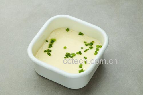 一位厨师正在厨房操作台前整理新鲜小葱,一部分整株放入装有清水的玻璃瓶中准备冷藏保存,另一部分已切碎撒在刚出锅的蒸蛋表面,旁边配有陶瓷调料罐与木制砧板,展现家庭健康烹饪场景 厨房中小葱的储存与使用场景