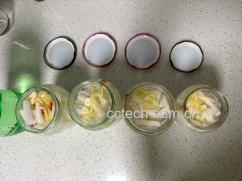 一个透明玻璃密封罐内盛放着用糖醋液腌制的紫色水果萝卜条,液体清澈微黄,萝卜条颜色鲜艳呈粉紫色,罐旁摆放着一双竹筷和一小碟辣椒面 玻璃罐中腌制的紫色水果萝卜条