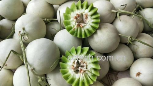 一个完整的玉菇甜瓜放置在木质砧板上,瓜体呈椭圆形,外皮为浅乳白色略带淡黄晕染,表面光滑无网纹,顶部保留新鲜绿色瓜蒂;切开一半展示内部透明水润的乳白色果肉,中心有少量黑色种子排列成环状,背景为自然光线下的厨房台面,突出其新鲜度与食用场景 玉菇甜瓜整体外观特写