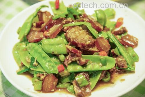 一位家庭主妇正在燃气灶前用铁锅翻炒青豌豆,锅中食材翻腾,可见绿色豆粒与橙色胡萝卜丁交织,动作流畅,厨房环境整洁,窗外透进自然光,营造温馨烹饪氛围 家庭厨房中正在翻炒青豌豆的场景