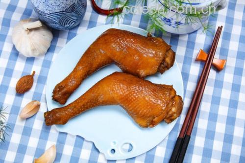 一只深盘中盛放两只色泽红亮的大鸡腿,周围环绕着浓郁酱汁,旁边点缀几段翠绿葱花,背景虚化的餐桌上配有米饭碗和筷子,展示诱人的家常美食氛围 红烧大鸡腿成品装盘