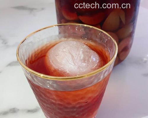 一瓶透明玻璃瓶中盛装着金红色的红梅果酒,酒液清澈透亮,瓶底有少量沉淀物,旁边摆放着两个小酒杯,一杯已倒满酒液,背景为木质餐桌和几颗干制红梅果,整体氛围温馨雅致 成品红梅果酒展示