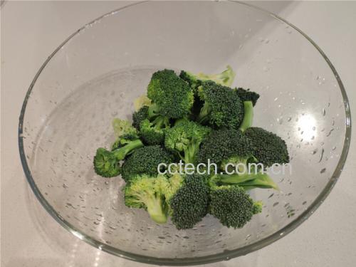 新鲜西兰花被放置在白色瓷盘中,旁边有一把不锈钢刀正在将其切成小朵,背景为淡木色料理台面,水中可见细小气泡,显示正在盐水中浸泡 西兰花的清洗与分切过程