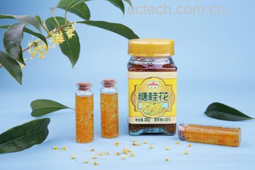 玻璃瓶装的淡黄色蜂蜜，标签标注“桂花风味蜂蜜”，旁边散落几朵干桂花