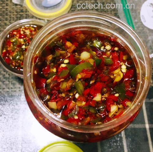 一瓶透明玻璃罐装的红褐色 homemade 辣椒酱摆在木桌上，旁边配有瓷勺和一碗冒着热气的白米饭