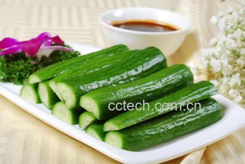一盘刚出锅的油焖小黄瓜盛放在白色瓷盘中,菜肴油光发亮,黄瓜段呈琥珀色,点缀着红色小米辣和蒜片,背景为简约中式餐桌